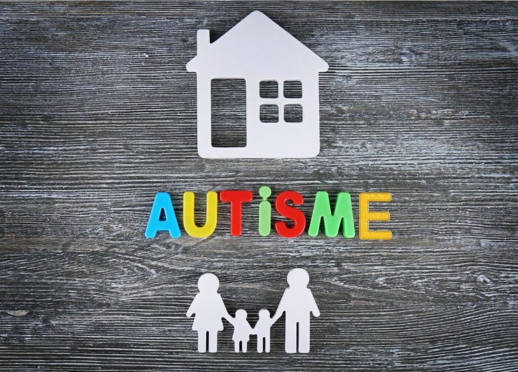 Sur une ardoise, des objets en plastique : une maison, dessous les lettres formant le mot "autisme" et encore dessus des silhouettes d'une famille : la maman et le papa encadrant 2 enfats et les tenant par la main