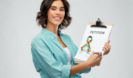 une dame tient un porte page où il est inscrit pétition et dessiné un ruban multicolore, symbole de l'autisme